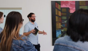 Brandon leads a tour. He is a white man with a beard and long brown hair pulled back, wearing a short sleeved blue shirt and holding a notebook in his hands that he gestures with while speaking to a group of college students.